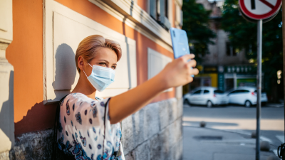 Nainen ottaa puhelimella selfietä seinän edessä kasvomaski naamalla.
